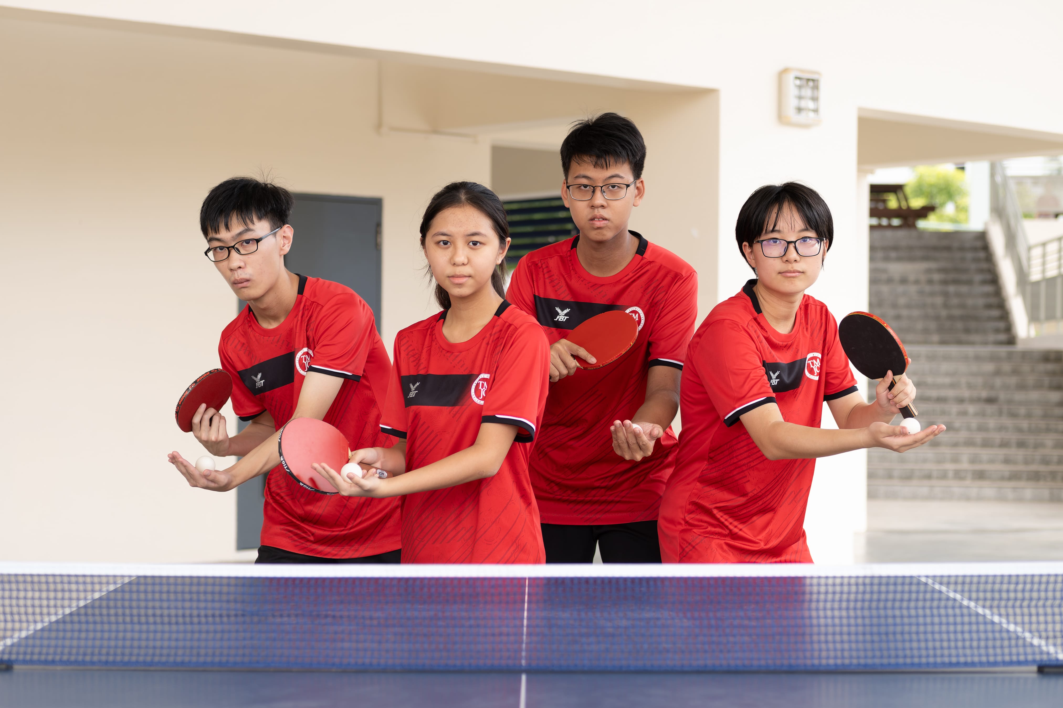 Four TMJC students in red jerseys pose at a table tennis table, each holding a paddle and ball in a serving stance.